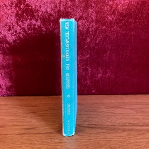 May include: A teal-colored book titled "New Testament Greek for Beginners" by MacMillan. The book is standing upright on a wooden surface, with a burgundy background. The book's spine is visible, displaying the title in white text.