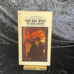 May include: A vintage paperback book titled "The Sea Wolf" by Jack London. The cover features an illustration of a man in a dark coat and cap, with a red and orange background. The book is propped up on a small wooden stand.