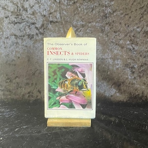 May include: A vintage book titled "The Observer's Book of Common Insects & Spiders" by E.F. Linssen & L. Hugh Newman. The cover features a close-up image of an insect on a pink flower. The book is displayed on a small, gold-colored stand.