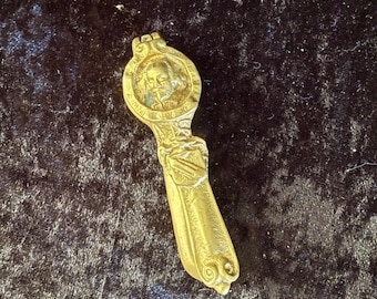 May include: A gold-colored nutcracker with a detailed design. The nutcracker features a round head with a profile of a man's face. The handle is shaped like a scroll and has intricate details.