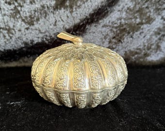 May include: A silver-colored, ornate trinket box with a pumpkin-like shape. The box features detailed floral patterns and a small handle on top. The box is sitting on a black surface.