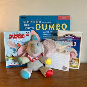 Könnte beinhalten: Eine Sammlung von Dumbo-Merchandise, darunter ein Plüschtier, ein Buch, eine Schallplatte, ein VHS-Band und ein Schreibwarensortiment. Das Plüschtier ist grau mit blauen, gelben und roten Akzenten. Das Buch trägt den Titel "Dumbo an Land, auf See, in der Luft". Die Schallplatte trägt den Titel "Shirley Temple erzählt die Geschichte von Walt Disneys Dumbo". Das VHS-Band trägt den Titel "Walt Disneys Meisterwerk-Sammlung Dumbo". Das Schreibwarensortiment umfasst ein Blatt Papier mit einem Dumbo-Motiv und einen Umschlag.