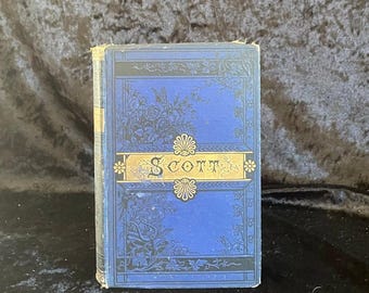 May include: A vintage book with a blue cover featuring floral and scrollwork designs. The title "Scott" is displayed in gold lettering on a gold banner. The book's edges show signs of wear, indicating its age.