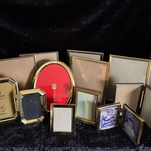 May include: A selection of gold-coloured picture frames in diverse shapes and sizes, including rectangular, oval, and octagonal. Some frames contain photos, while others are empty. The frames are arranged on a dark, textured surface.