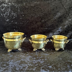 May include: A set of three graduated brass pots, each featuring a polished metallic finish, two side handles, and three ornate feet. The pots are arranged in a row, decreasing in size from left to right, showcasing a consistent design.