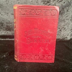 Peut inclure: Un livre rouge vintage intitulé "Safe Methods of Business". La couverture présente des motifs floraux en relief et le titre en lettrage élégant. Le dos affiche du texte supplémentaire. Le livre semble bien conservé.