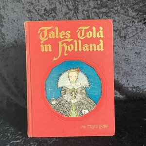 Puede incluir: Un libro antiguo titulado "Tales Told in Holland" con una portada roja. La portada presenta letras doradas y una ilustración de una mujer con un vestido histórico dentro de un marco azul y rojo. El libro es de "My Travelship".