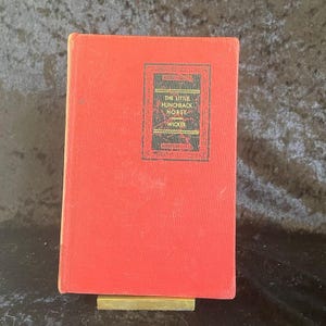 May include: A vintage book titled "The Little Hunchback Horse" by Wicker. The book has a bright red cover with a rectangular design in black and gold. The book is propped up on a small brass stand.