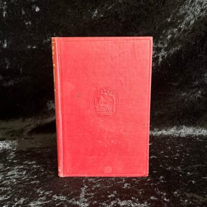Può includere: Un libro d'epoca con copertina rossa. La copertina presenta un'illustrazione in rilievo all'interno di una cornice circolare. Il dorso del libro è visibile e i bordi delle pagine mostrano usura. Il libro è in piedi.