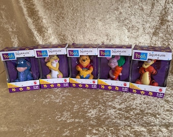 May include: Five Winnie the Pooh character bath squeeze toys in their individual packaging. Each box features a different character: Eeyore, Tigger, Piglet, Rabbit, and Winnie the Pooh. The boxes are yellow, purple, and orange with the words "Pooh Squeezie" printed on them.