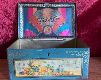 Könnte beinhalten: Eine vintage blaue Metalldose mit goldener Verzierung und einem Blumendesign auf der Vorderseite. Der Deckel hat ein farbenfrohes Design mit dem Text "Thornes" in einer dekorativen Schrift.
