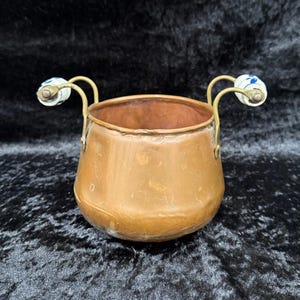 Może przedstawiać: Miedziany metalowy garnek z dwoma zakrzywionymi uchwytami. Uchwyty mają białe ceramiczne końcówki z niebieskimi kwiatowymi wzorami. Garnek ma zaokrągloną podstawę i szeroki otwór. Garnek jest prawdopodobnie zabytkowy lub antyczny.