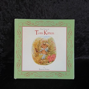 May include: The Tale of Tom Kitten by Beatrix Potter, a green book cover with a decorative border and an illustration of a kitten holding a smaller kitten.