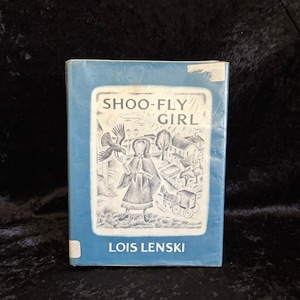 May include: A blue hardcover book titled Shoo-Fly Girl by Lois Lenski. The front cover features a white rectangular illustration of a person standing with a bird, a horse-drawn carriage, and houses. There is a small piece of tape on the top right corner.
