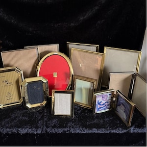 May include: A collection of various gold-toned picture frames in different shapes and sizes. Some frames are rectangular, others oval or octagonal. One frame features the text "Solid Brass". Several frames contain photos.