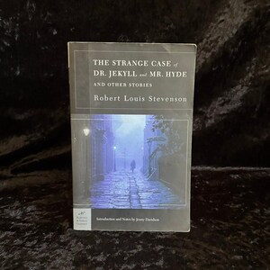 Peut inclure: Un livre de poche intitulé The Strange Case of Dr. Jekyll and Mr. Hyde and Other Stories de Robert Louis Stevenson. La couverture présente une illustration sombre et atmosphérique d'une rue pavée la nuit avec une silhouette solitaire.