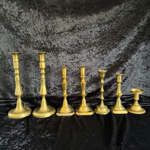 May include: A collection of seven antique brass candlesticks of varying heights and designs. The candlesticks have a warm, golden hue and feature detailed bases and stems. The arrangement showcases a range of styles, from tall, slender forms to shorter, wider ones.