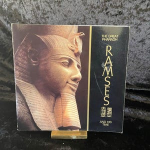May include: A book titled "Ramses and His Time" featuring a portrait of a pharaoh. The cover has a black background with gold lettering. The image shows a bust of the pharaoh in shades of brown and tan.