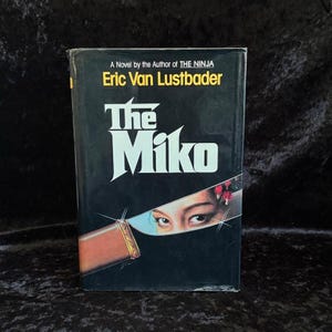 Puede incluir: Un libro de tapa dura titulado "The Miko" de Eric Van Lustbader. La portada negra presenta el título en blanco y los ojos de una mujer vistos a través de una cuchilla. El autor es también el de "The Ninja".