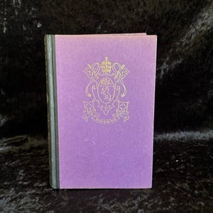 May include: A hardcover book with a purple fabric cover and a dark green spine. The front cover features a gold embossed emblem depicting a crest with a bear, a bird, and a papal tiara, with the Latin text EX ORIENTE LUX inscribed below.