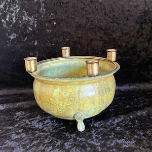 May include: A round, brass-toned metal bowl with a weathered, greenish patina. The bowl features three small, shell-shaped feet at the base and four cylindrical candle holders attached to the upper rim, evenly spaced around the circumference.
