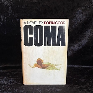 Può includere: Un libro tascabile vintage intitolato "COMA" di Robin Cook. La copertina presenta il titolo in grassetto nero e un'illustrazione di una persona. Il libro ha uno sfondo color crema.
