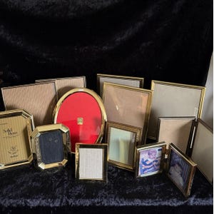 May include: A collection of various gold-toned picture frames in different shapes and sizes. Some frames are rectangular, others oval, and some have ornate detailing. One frame is labeled "Solid Brass". The frames are arranged on a dark, textured surface.