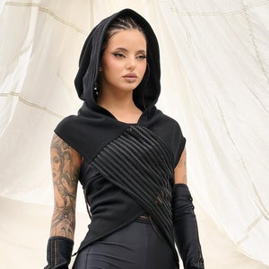 Puede incluir: Una mujer con una capucha negra con un diseño de cruz, pantalones cortos negros y guantes negros sin dedos. Lleva botas negras con hebillas plateadas.