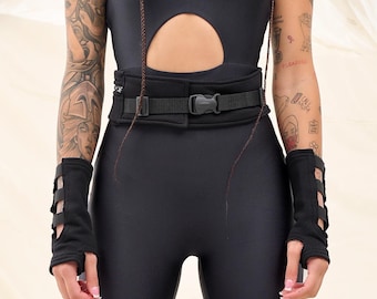 May include: A woman wearing a black jumpsuit with a cut-out design and a belt. She has black braids with a criss-cross design and multiple tattoos on her arms. The background is a white fabric.