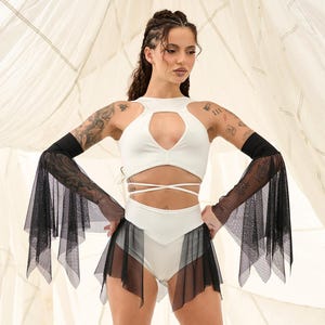 May include: A white crop top with a keyhole cutout and matching high-waisted shorts. The outfit is paired with black mesh arm sleeves and a black mesh skirt. The model is wearing black platform boots.