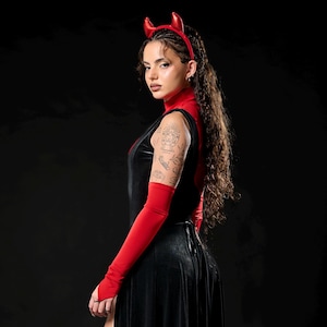 May include: A black velvet dress with a high slit, paired with red arm warmers and a red turtleneck. The outfit is accessorized with a red devil horn headband and platform boots, creating a gothic or costume aesthetic.