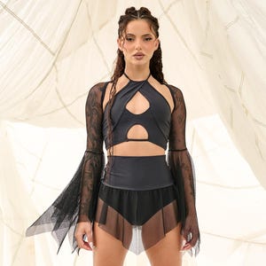 May include: A black two-piece outfit. The halter top has cutouts, and the high-waisted shorts have a sheer skirt overlay. The outfit includes long, sheer sleeves and platform boots. The background is white fabric.