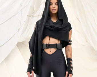 May include: A person wearing a black hooded top with a black belt and black trousers with straps and buckles. The person is standing in front of a white fabric background.