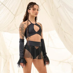 May include: A black halter top with a keyhole cutout, matching arm sleeves, and a mesh skirt. The outfit is paired with platform boots. The background is a white fabric.