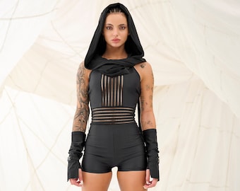 May include: A woman wearing a black hooded jumpsuit with a mesh panel and black boots. She is standing in front of a white backdrop.