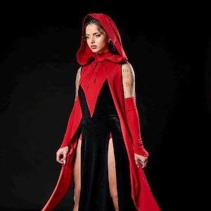 May include: A black velvet gown with high slits and a red hooded cape. The cape has a deep V-neckline and long sleeves. The model is wearing black platform boots. The outfit is set against a black background.