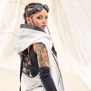 K&ouml;nnte beinhalten: Ein futuristisches Outfit mit einer wei&szlig;en Kapuzenweste und schwarzen Details, kombiniert mit schwarzen Leggings und langen schwarzen &Auml;rmeln. Das Model tr&auml;gt eine schwarze Brille und hat ein Tattoo auf dem Arm. Das Outfit hat einen postapokalyptischen Stil.