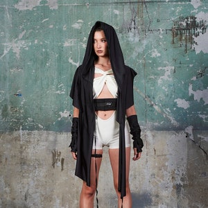 May include: A person wearing a black hooded top with a white bodysuit and black boots. The person is standing in front of a green and grey textured wall.