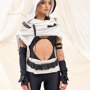 K&ouml;nnte beinhalten: Ein futuristisches Outfit mit einem wei&szlig;en Kapuzenmantel mit schwarzem Besatz, kombiniert mit einem schwarzen Body und Leggings. Das Ensemble umfasst einen breiten G&uuml;rtel mit Schnalle, Armstulpen und Beinschlaufen. Die Gesamt&auml;sthetik ist modern und kantig.