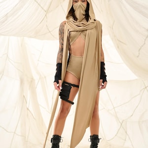 May include: A tan-colored hooded outfit with a face covering and long scarf. The outfit includes a crop top, high-waisted shorts, and black combat boots. Black arm and leg accessories complete the look.