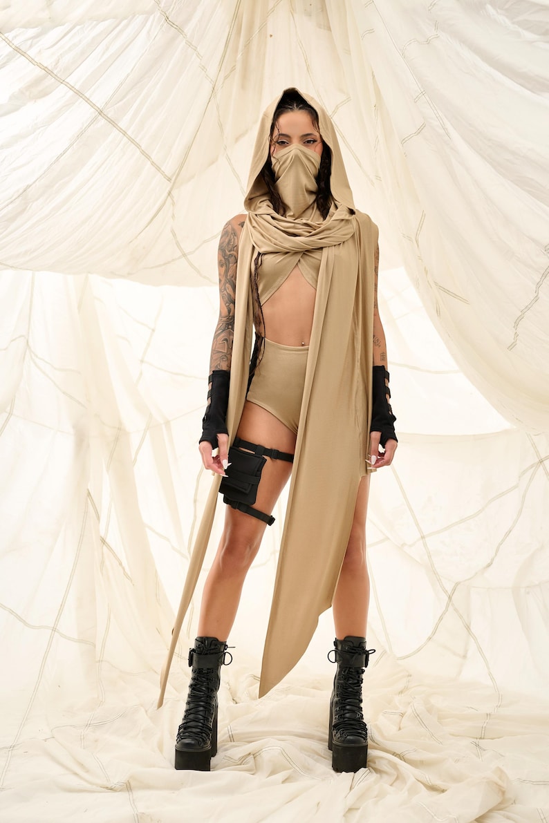 May include: A tan-colored hooded outfit with a face covering and long scarf. The outfit includes a crop top, high-waisted shorts, and black combat boots. Black arm and leg accessories complete the look.