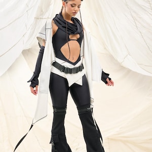 K&ouml;nnte beinhalten: Ein futuristisch anmutendes Outfit mit einem schwarzen Body mit Ausschnitt, kombiniert mit schwarzen Leggings und Schlaghosen. Das Ensemble umfasst eine wei&szlig;e Jacke, einen schwarzen G&uuml;rtel und eine Brille. Der Hintergrund ist neutral.