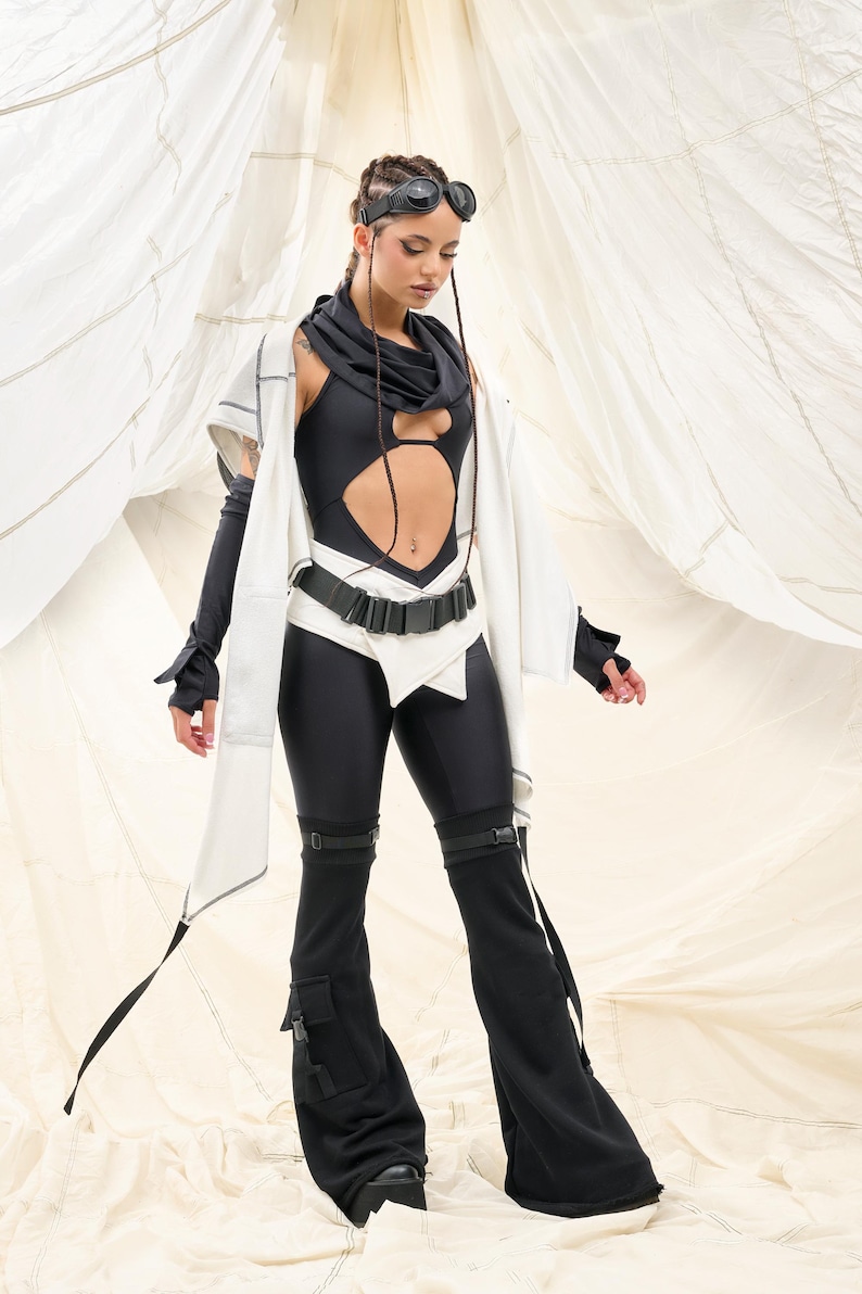 K&ouml;nnte beinhalten: Ein futuristisch anmutendes Outfit mit einem schwarzen Body mit Ausschnitt, kombiniert mit schwarzen Leggings und Schlaghosen. Das Ensemble umfasst eine wei&szlig;e Jacke, einen schwarzen G&uuml;rtel und eine Brille. Der Hintergrund ist neutral.