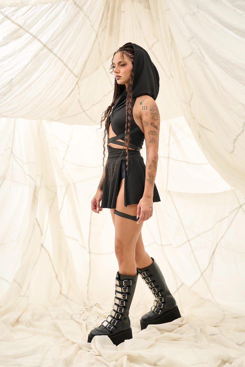 Pode incluir: Um conjunto preto de top cropped com capuz e saia com design de embrulho. O traje &eacute; combinado com botas de plataforma at&eacute; o joelho com v&aacute;rias fivelas prateadas. A modelo est&aacute; em frente a um fundo branco.