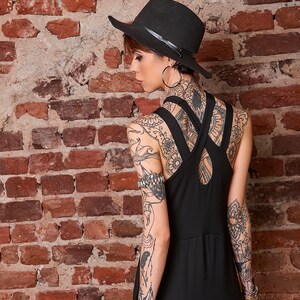 May include: A woman wearing a black fedora hat and a black sleeveless dress with a criss-cross back detail. She has intricate tattoos on her arms and back.