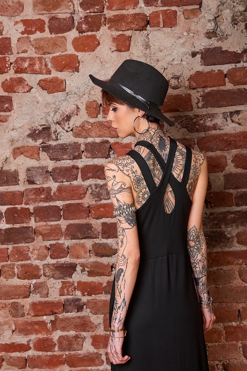 May include: A woman wearing a black fedora hat and a black sleeveless dress with a criss-cross back detail. She has intricate tattoos on her arms and back.