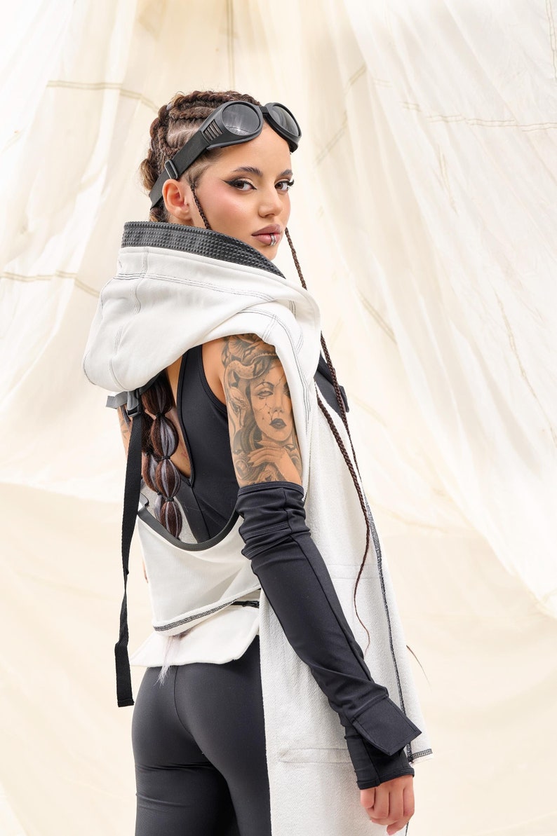 K&ouml;nnte beinhalten: Ein futuristisches Outfit mit einer wei&szlig;en Kapuzenweste und schwarzen Details, kombiniert mit schwarzen Leggings und langen schwarzen &Auml;rmeln. Das Model tr&auml;gt eine schwarze Brille und hat ein Tattoo auf dem Arm. Das Outfit hat einen postapokalyptischen Stil.