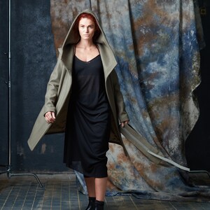 Puede incluir: Una mujer con el pelo rojo lleva un abrigo largo de color verde oliva con capucha sobre un vestido negro. Est&aacute; de pie frente a un fondo estampado azul y marr&oacute;n.