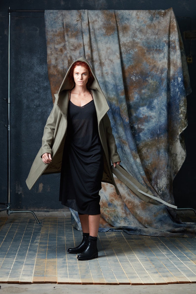 Puede incluir: Una mujer con el pelo rojo lleva un abrigo largo de color verde oliva con capucha sobre un vestido negro. Est&aacute; de pie frente a un fondo estampado azul y marr&oacute;n.