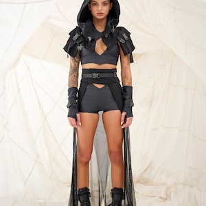 以下が含まれることがあります： 黒いフード付きトップス、黒いベルト、黒いショートパンツを着用した女性。黒いブーツと黒いシースルーのスカートを履いています。背景は白い布です。